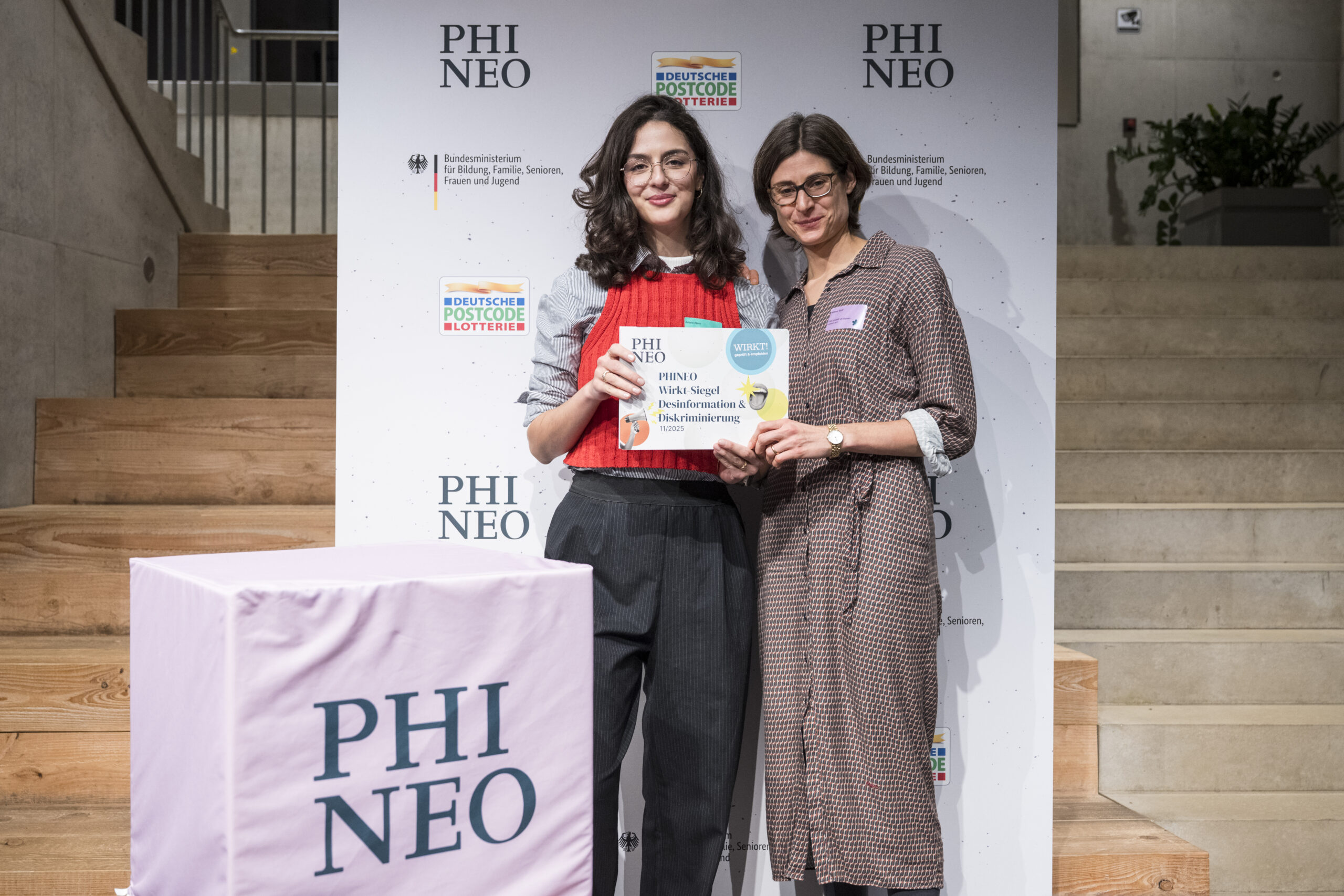Ariane Alam und Helene Wolf von FAIR SHARE stehen nebeneinander und halten gemeinsam eine Urkunde des „PHINEO Wirkt-Siegels“ hoch. Ariane Alam trägt ein rotes Oberteil mit grauem Hemd, Helene Wolf ein gemustertes Kleid. Beide lächeln in die Kamera. Im Hintergrund ist eine weiße Wand mit PHINEO-Logos sowie Logos des Bundesministeriums und der Deutschen Postcode Lotterie zu sehen. Links im Vordergrund steht ein würfelförmiges Podest mit der Aufschrift „PHINEO“.